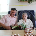 Gratulant u oslavy 100. narozenin uživatelky, květina v ruce, koláčky